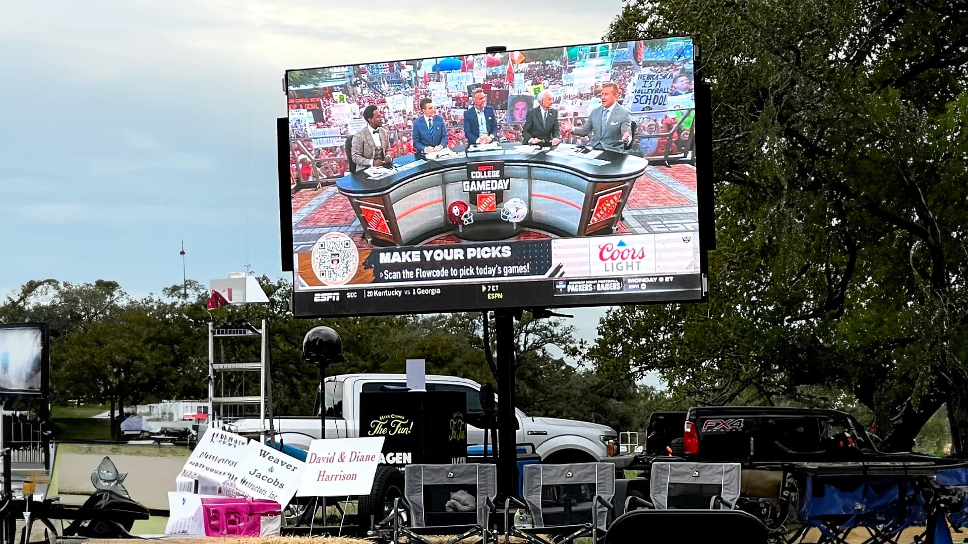 Crowd tailgating around a mobile LED screen showing a college football broadcast